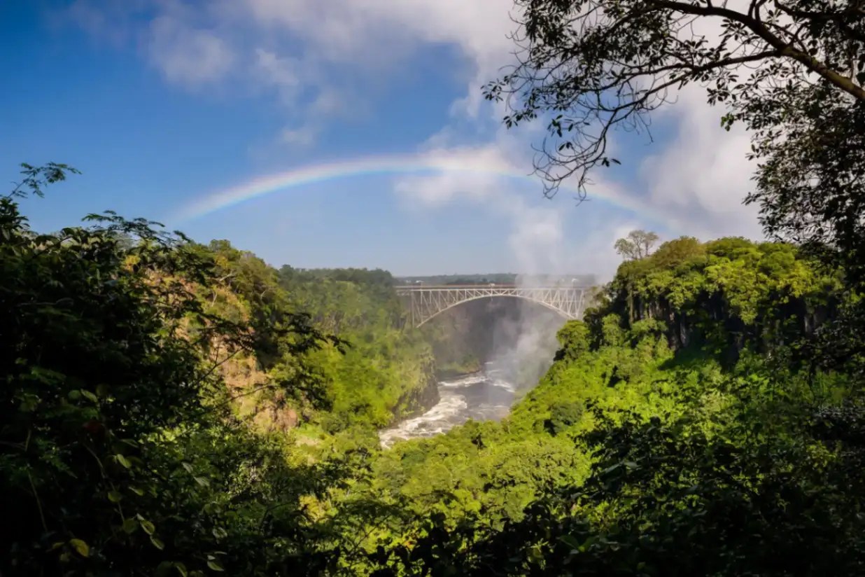 A scenic view of the Livingstone Bridge arching over the Zambezi River with a vibrant rainbow rising above Victoria Falls.